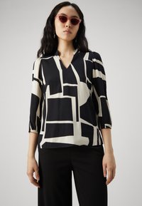 Blusa a fantasia bianca e nera con scollatura a V, maniche a tre quarti e polsini elasticizzati. Tessuto morbido con stampe geometriche.