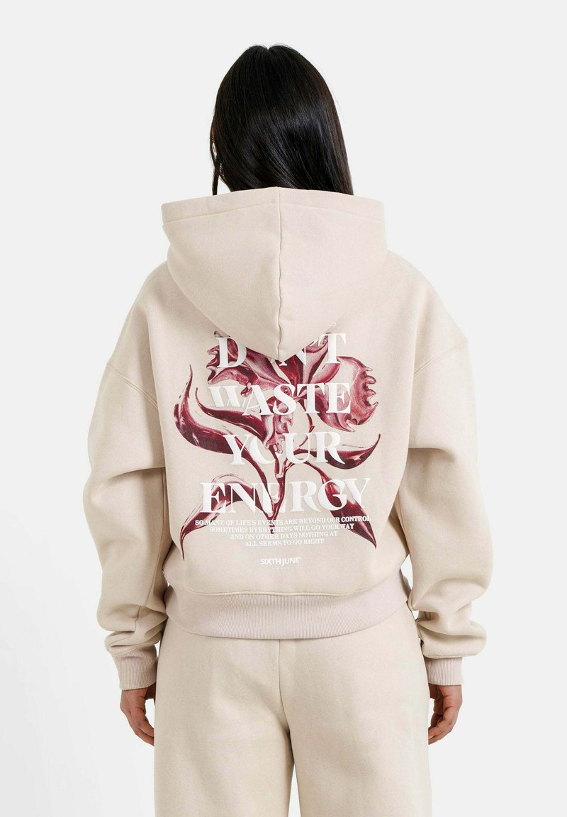Felpa beige con una grande grafica in rosso e testo bianco sul retro. Presenta un cappuccio e polsini a costine, realizzata in materiale morbido.