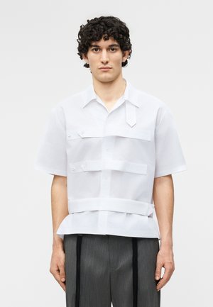 Jeune homme aux cheveux bouclés portant une chemise blanche à manches courtes avec plusieurs lanières horizontales et un pantalon gris à fines rayures.