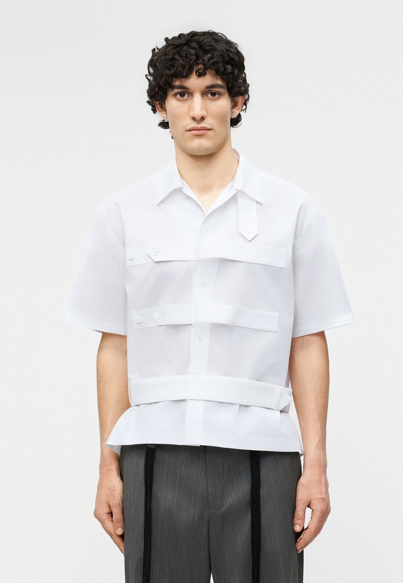 Νεαρός με κατσαρά μαλλιά που φοράει λευκό πουκάμισο με κοντά μανίκια και κουμπιά, με πολλαπλές οριζόντιες τιράντες, και γκρι παντελόνι με λεπτές ρίγες.