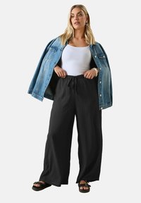 Jeansowa kurtka, biała koszulka na ramiączkach i czarne szerokie spodnie o strukturalnym wykończeniu. Czarne sandały i srebrna bransoletka dopełniają strój.