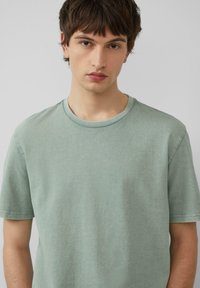 Jasnozielona koszulka z krótkim rękawem, wykonana z miękkiej bawełny, z okrągłym dekoltem i gładką fakturą, charakteryzująca się minimalnymi szwami i luźnym krojem.