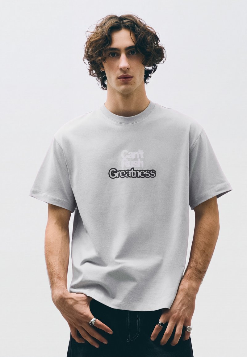 Jeune homme aux cheveux bruns bouclés porte un t-shirt gris clair avec le texte "Can't Rush Greatness" et a des bagues aux deux mains.