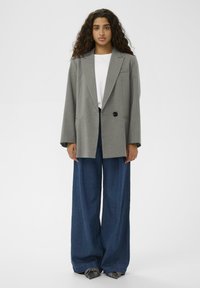 Femeie cu păr lung și creț, îmbrăcată într-un sacou gri cu dungi, cămașă albă, blugi largi de culoare albastră și pantofi gri cu vârf ascuțit, stând în fața unui fundal alb.