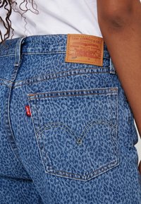 Blå denimjeans med en texturerad yta, med en brun läderpatch och en röd logotagg på bakfickan. Utmärkande sömnadsdesign.