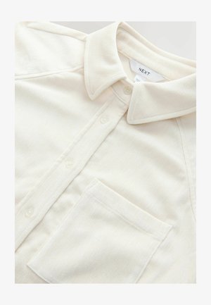 Krém színű, galléros ing, puha anyagból készült, mellzsebbe, gombos záródással és finom textúrával.