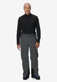 Graue wasserdichte Skihosen mit verstellbaren Trägern, kombiniert mit einem schwarzen Langarmoberteil. Merkmale sind flache Nähte und Skischuhe mit Schnallen.