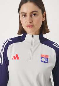 Chaqueta deportiva blanca y morada con collar alto, cierre de cremallera y logotipo rojo de Adidas. Presenta el escudo del Olympique de Lyon en el pecho.