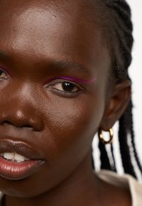 Gros plan sur un visage à la peau foncée, avec des cheveux tressés et un eyeliner magenta audacieux et courbé. Boucles d'oreilles en or dans l'oreille. Texture de peau lisse.