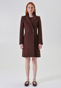 Vrouw met rood haar draagt een knielange bruine double-breasted jas en ballerina's met luipaardprint, staande tegen een effen grijze achtergrond.