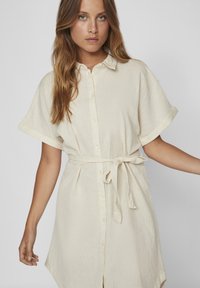 Svijetlo bež haljina košulja od lana, s ovratnikom, kratkim zavrtnutim rukavima, prednjim gumbima i vezanjem u struku za strukturirani kroj.