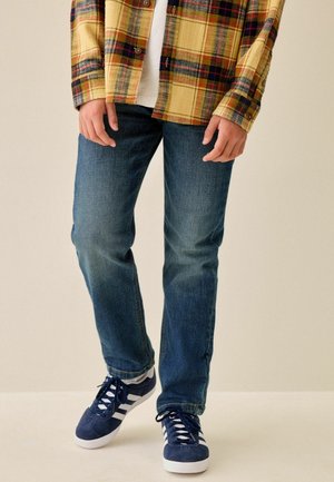 Personne portant un jean bleu, des baskets bleu marine et blanches, et une chemise à carreaux jaune et rouge, les mains détendues le long du corps.
