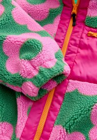 Jachetă din fleece cu bază verde, modele mari florale roz, fermoar portocaliu aprins și finisaj moale, texturat.