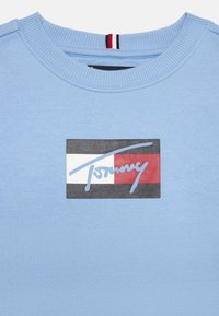 Hellblaues Sweatshirt mit einem rechteckigen Logo-Patch in rot, weiß und schwarz sowie einer blauen Unterschrift. Glatte Textur, runder Ausschnitt.