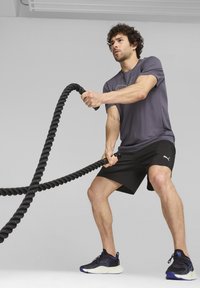 Fekete sportnadrág és egy szürke póló grafikai mintával. A személy egy vastag fekete harci kötelet fog, és fekete sportcipőt visel, amelyen lila kiegészítők találhatók.