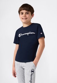 Temno modra bombažna majica s belim logotipom "Champion" spredaj, oblečena z sivimi trenirkami, ki imajo temen logotip ob strani.