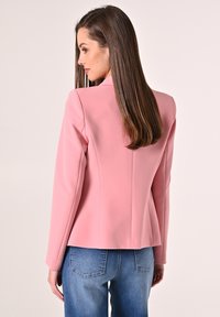 Rosa Blazer mit einer taillierten Passform, glatter Textur und leichtem Glanz, kombiniert mit blauen Jeans. Verfügt über einen maßgeschneiderten Kragen und klare Linien.