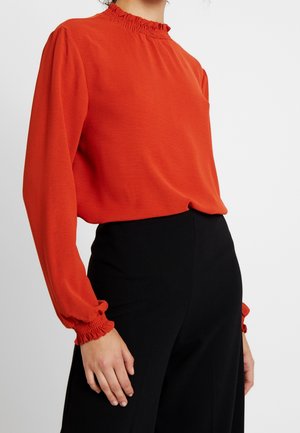 Femme portant un chemisier rouge à manches longues avec col et poignets volantés, associé à un pantalon noir taille haute sur fond blanc.