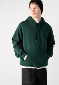 Sudadera con capucha en verde oscuro, hecha de tela suave, cuenta con un bolsillo frontal y cordón, usada sobre una camisa blanca y pantalones oscuros.