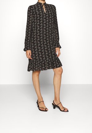 Femme portant une robe noire à manches longues avec un motif floral et "MC", ourlet à volants, encolure en trou de serrure, assortie à des sandales à talons noires à brides.