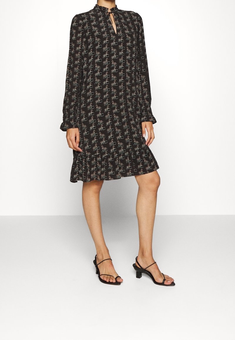 Femme portant une robe noire à manches longues avec un motif floral et "MC", ourlet à volants, encolure en trou de serrure, assortie à des sandales à talons noires à brides.
