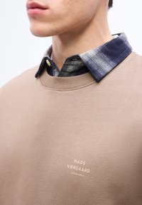Homme portant un sweat-shirt beige par-dessus une chemise à carreaux bleue et grise, affichant le logo du sweat-shirt "Mads Nørgaard Copenhagen" sur la poitrine.