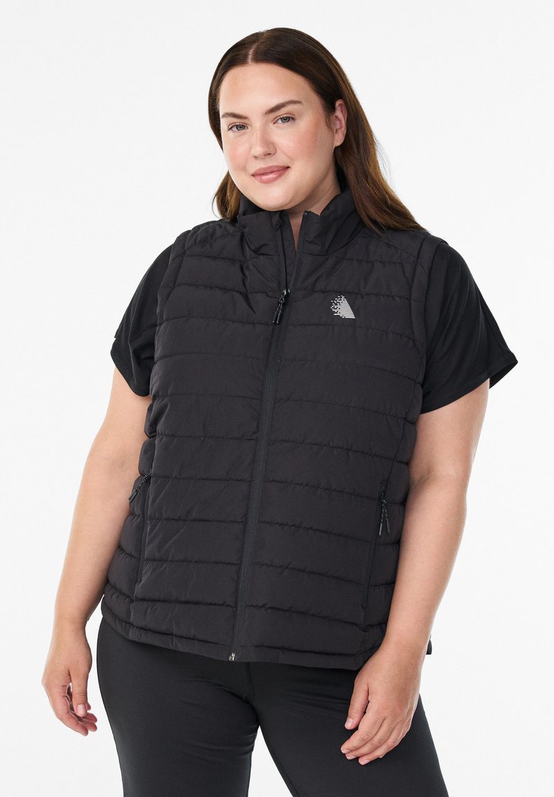 Frau in einer schwarzen gesteppten ärmellosen Weste über einem schwarzen kurzärmeligen Shirt, steht mit einem leichten Lächeln vor einem weißen Hintergrund.
