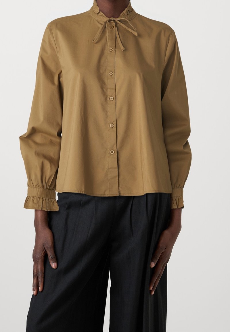 Femme portant un chemisier manches longues couleur moutarde avec des boutons à l’avant et un col volanté, associé à un pantalon noir ample.
