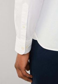 White long-sleeve shirt with buttoned cuff, featuring a smooth texture and rounded hem. The fabric appears lightweight and slightly wrinkled.