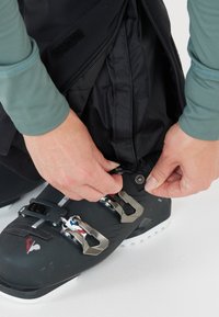 Zwarte skibroek met ritsen en drukkers, die wordt aangepast door een hand boven een zwarte skiboot met metalen gespen. De achtergrond is wit.