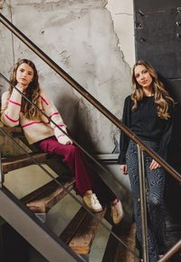 Twee modellen op houten trappen. De een draagt een crèmekleurige trui met roze strepen en bordeauxrode broek; de ander heeft een zwarte textuurtop en een broek met patroon.