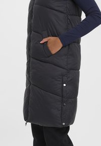 Fodrad svart väst med chevron-quiltat mönster, sidofickor och tryckknappsdetaljer. Har dragkedjestängning och hög krage för värme.