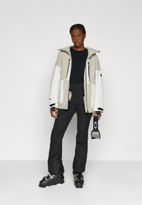 Hiihtotakki vaaleanbeige ja valkoinen, jossa on huppu, vetoketjulliset taskut ja säädettävät hihansuut. Yhdistetty mustiin housuihin ja hiihtokenkään.