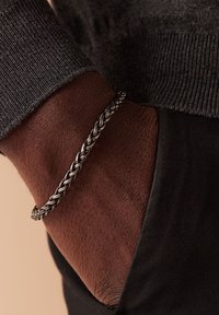 Zilveren gevlochten armband om een donkere huidpols, gecombineerd met een donkere trui, die textuur en een intrigerend weefontwerp laat zien.