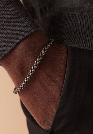 Zilveren gevlochten armband om een donkere huidpols, gecombineerd met een donkere trui, die textuur en een intrigerend weefontwerp laat zien.
