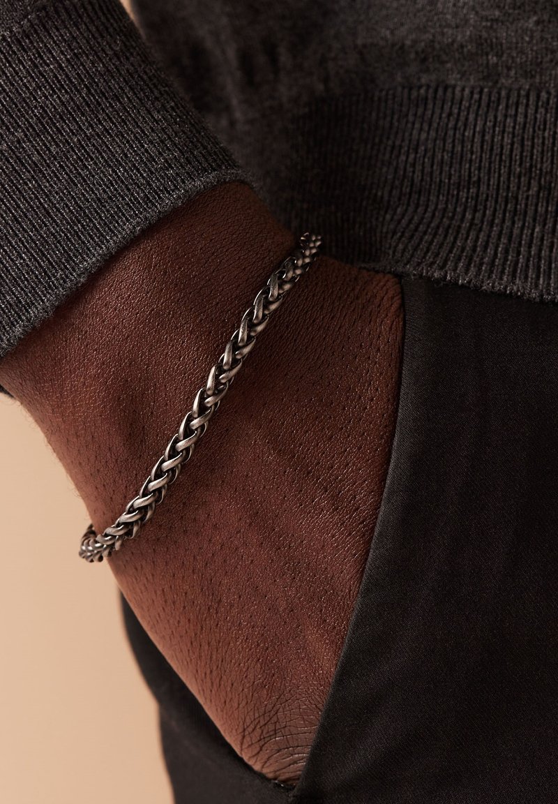 Zilveren gevlochten armband om een donkere huidpols, gecombineerd met een donkere trui, die textuur en een intrigerend weefontwerp laat zien.