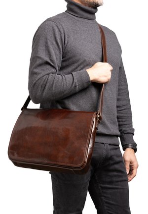 Man draagt een grijze coltrui en zwarte broek, en houdt met zijn linkerhand een bruine leren boodschappentas over zijn schouder.