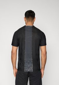 Μαύρο αθλητικό πουκάμισο με υφή, διαθέτει κεντρικό σκούρο πάνελ και ανακλαστικές λεπτομέρειες. Κοντά μανίκια και στρογγυλή λαιμόκοψη.