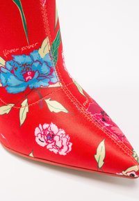Bottine en tissu rouge avec un bout pointu, motifs floraux en bleu et rose, avec des feuilles vertes, et détails de couture visibles.