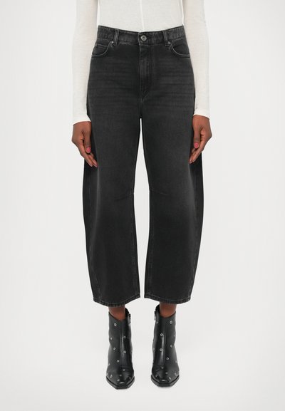 Hochtaillierte schwarze Denim-Jeans mit weitem Bein, ausgestattet mit Vorderaschen und einem klassischen Knopfverschluss. Kombiniert mit schwarzen Knöchelschuhen mit Ösen.