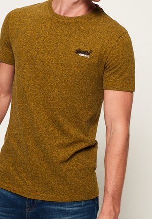 Mann trägt ein eng anliegendes, kurzärmeliges, senfgelbes, strukturiertes T-Shirt mit einem kleinen schwarz-weißen Logo auf der linken Brust.