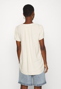 Ljus beige kortärmad topp med rund halsringning, texturerad tyg, veckad detalj på baksidan och en längre nederkant. Matchad med denimshorts.