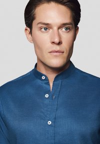 Niebieska lniana koszula z kołnierzykiem typu stójka, z przodu zapinana na guziki w kontrastującym białym kolorze. Gładka tekstura o swobodnym designie.