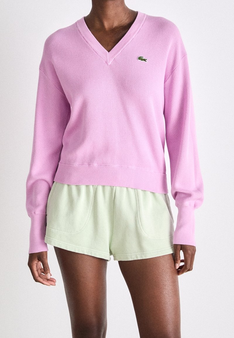 Persona vestida con un suéter V de color rosa claro con el logo de Lacoste y pantalones cortos casuales de color verde claro, de pie frente a un fondo liso.