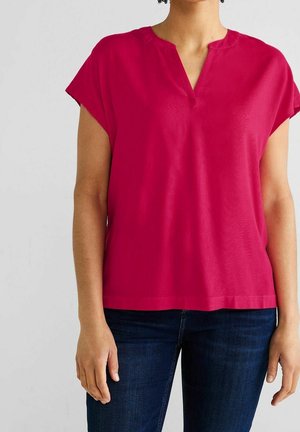 Henkilö, jolla on lyhythihainen, kirkkaan vaaleanpunainen V-kaula-aukollinen toppi ja tummansiniset farkut, seisomassa yksiväristä vaaleaa taustaa vasten.