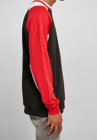 Persona con una sudadera negra de mangas rojas y detalles blancos, combinada con unos pantalones vaqueros gris claro, posando de perfil contra un fondo liso.