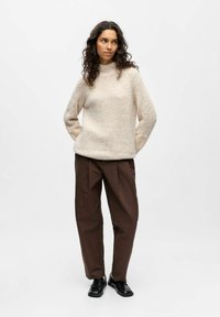 Krēmkrāsas vilnas džemperis ar augstu apkakli, teksturētu korpusu un dekoratīvām mežģīņu adījuma detaļām uz piedurknēm, kopā ar tumši brūnām biksēm un melniem apaviem.