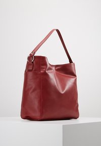 Sac cabas en similicuir rouge à texture lisse, forme souple, avec une seule bandoulière réglable. Doté d'un détail anneau en métal argenté.