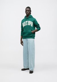 Zöld kapucnis pulóver "GRANT 1949" fehér felirattal, világoskék bő szárú farmerral és fekete cipővel párosítva. Sima anyag és laza szabás.