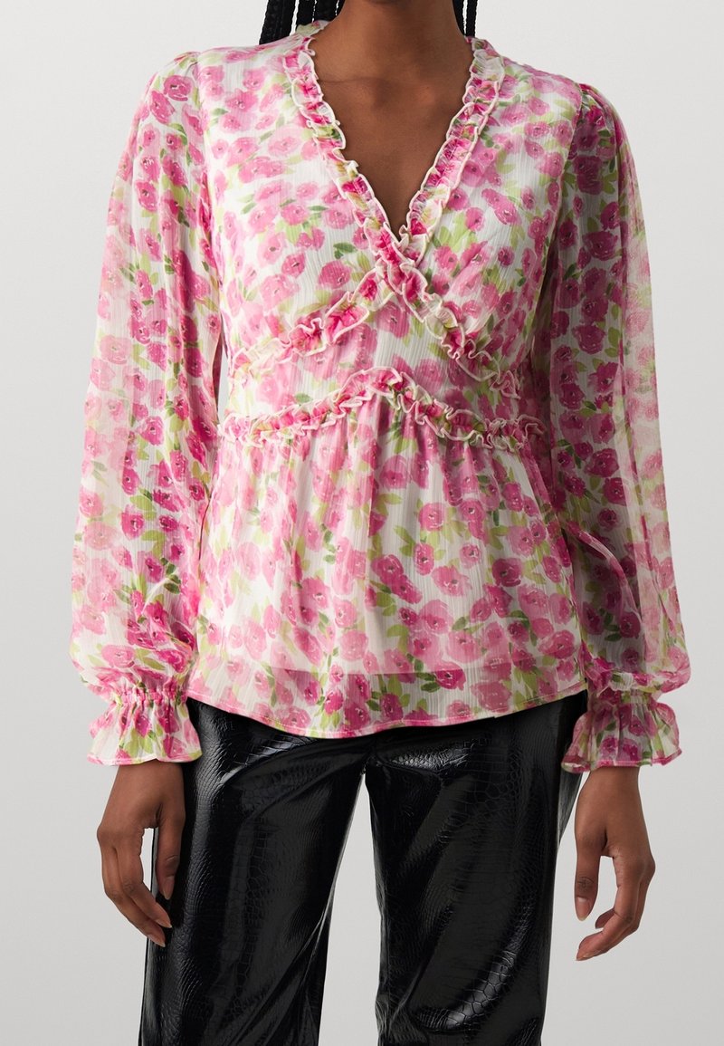 Femme portant un chemisier rose à volants avec des fleurs, manches longues, et un pantalon noir texturé, sur fond uni.